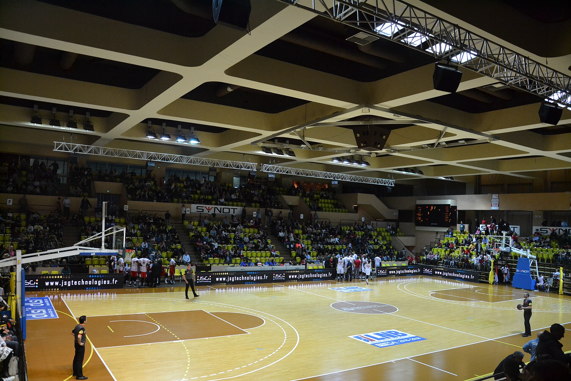 A basketball arena during a match, seen from high in the stand: a pale wooden court marked with the restricted area, free-throw circle and three-point line, backboards at both ends and tiered seating around it.