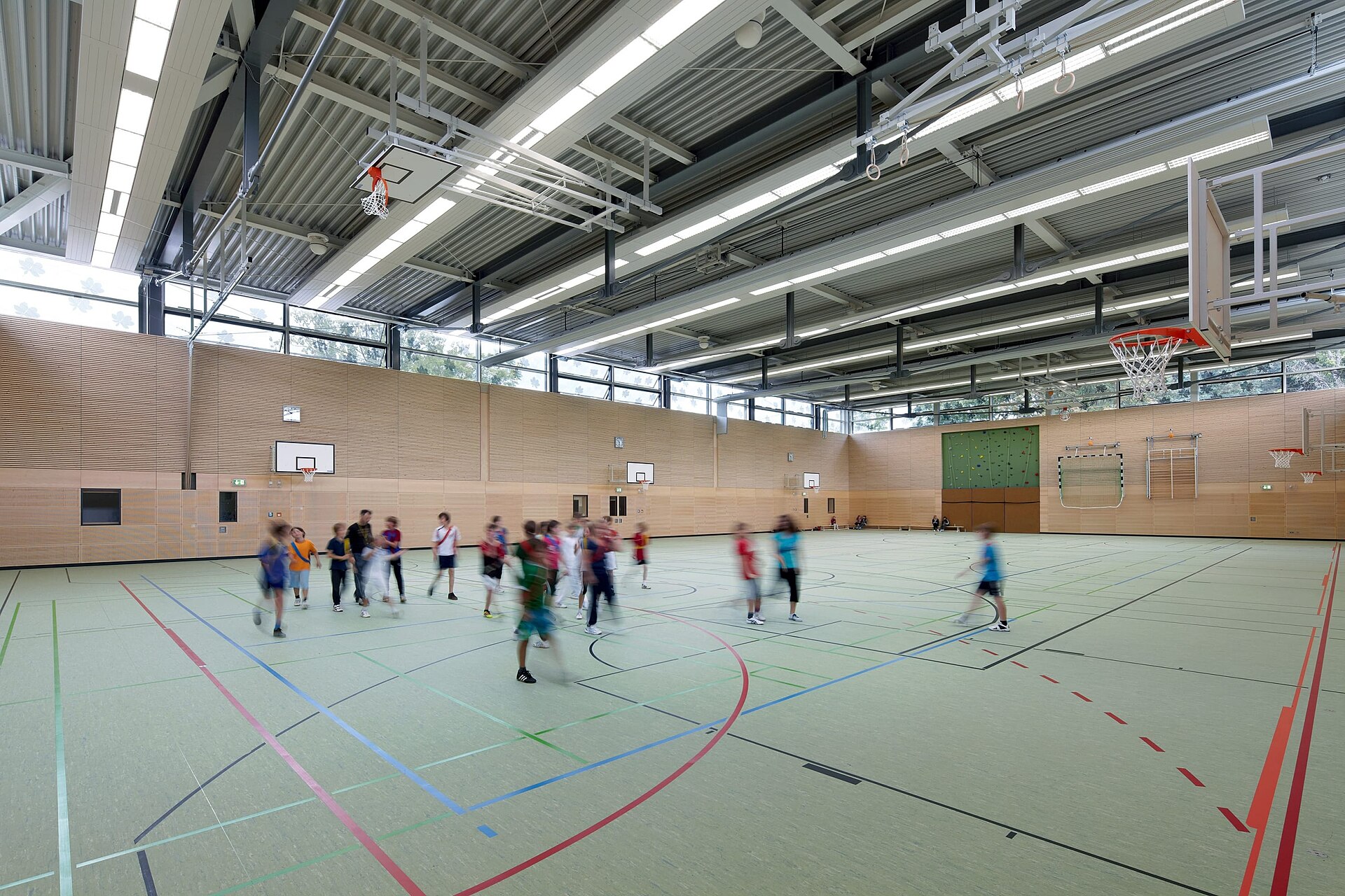 The interior of a modern indoor sports hall with a pale wooden floor and multiple sets of coloured court markings.