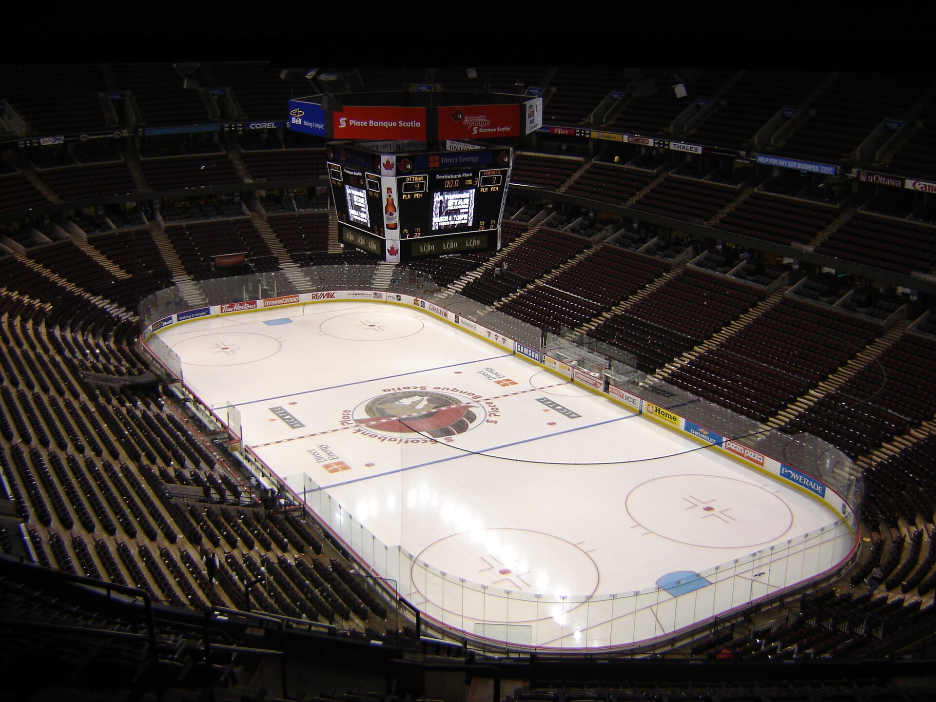 The interior of an empty indoor ice hockey arena, the white sheet marked with lines and face-off circles, seating rising on all sides.