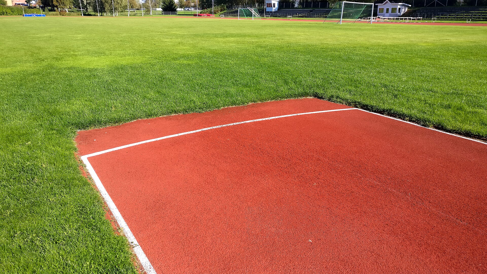 The end of a red synthetic javelin runway marked with a curved white throwing arc, opening onto a mown grass landing sector, with goalposts and a small stadium building in the distance.