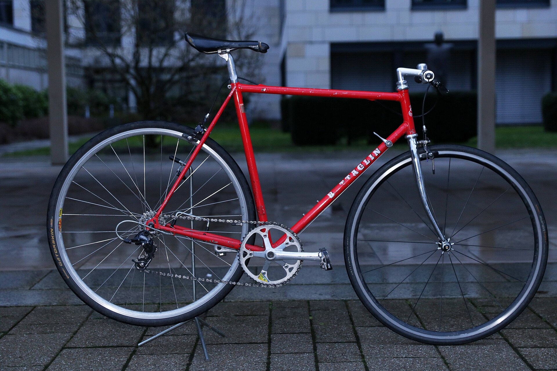 A red steel road bicycle on a stand, photographed side-on in low evening light, its diamond frame, chainset and shallow box-section rims filling the frame.