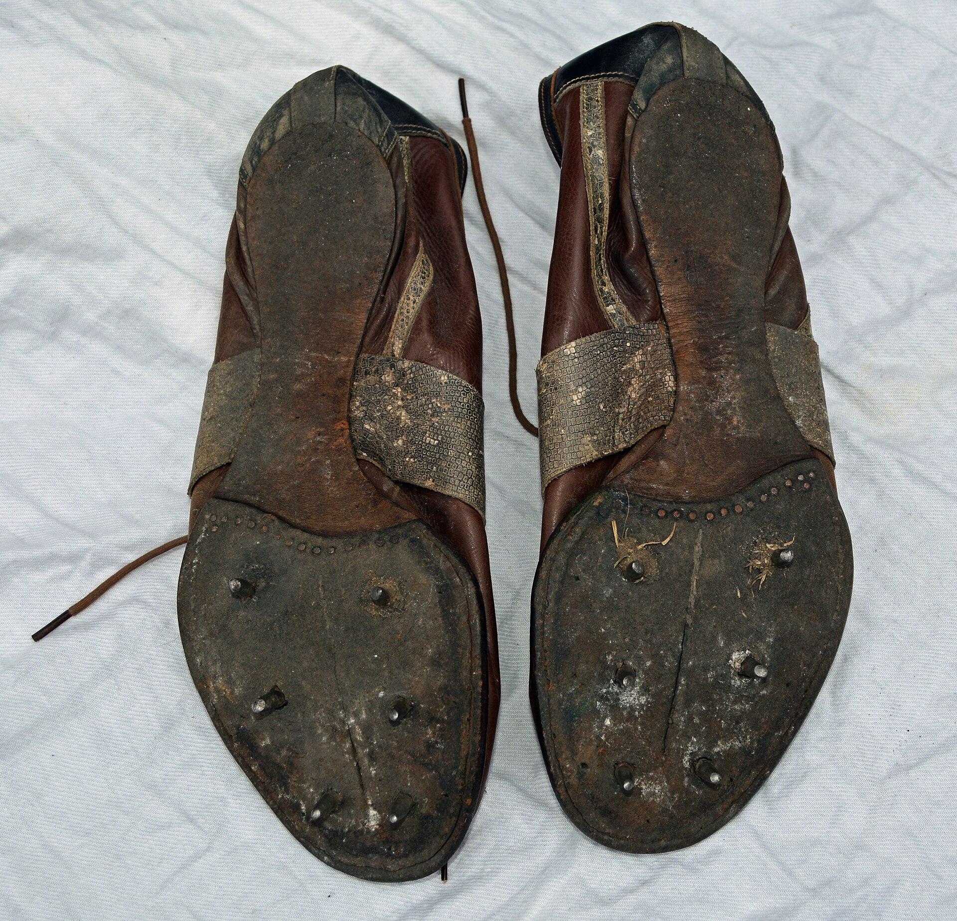 A worn pair of old brown leather running spikes lying sole-upward on white cloth, metal studs set into each thin flat forefoot plate and no studs under the heels.