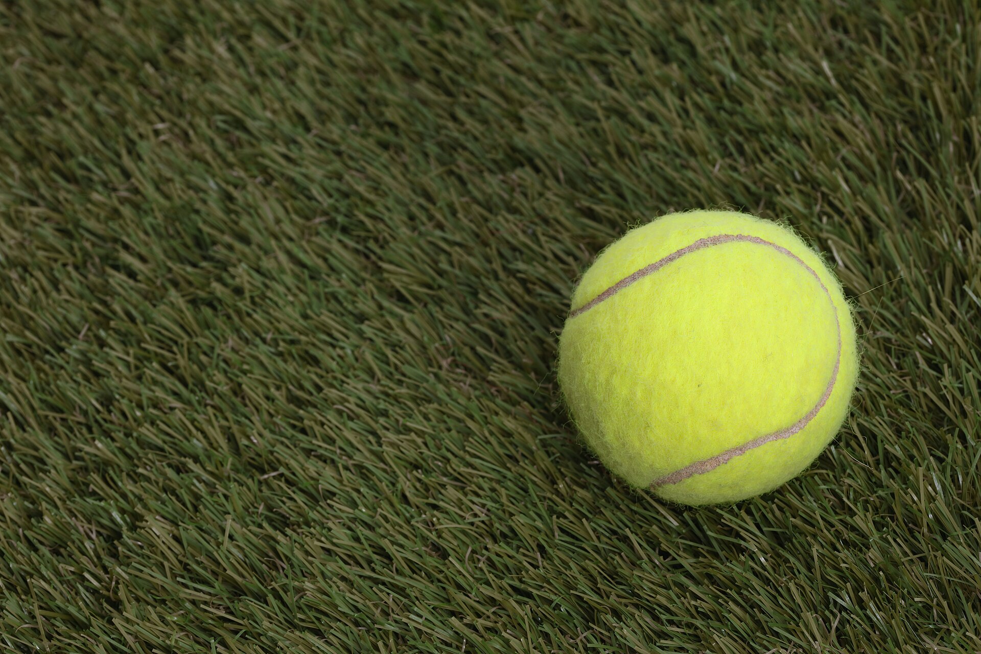 A single yellow tennis ball resting on artificial grass, its felt nap and pale curving seam sharp against the blurred synthetic fibres around it.