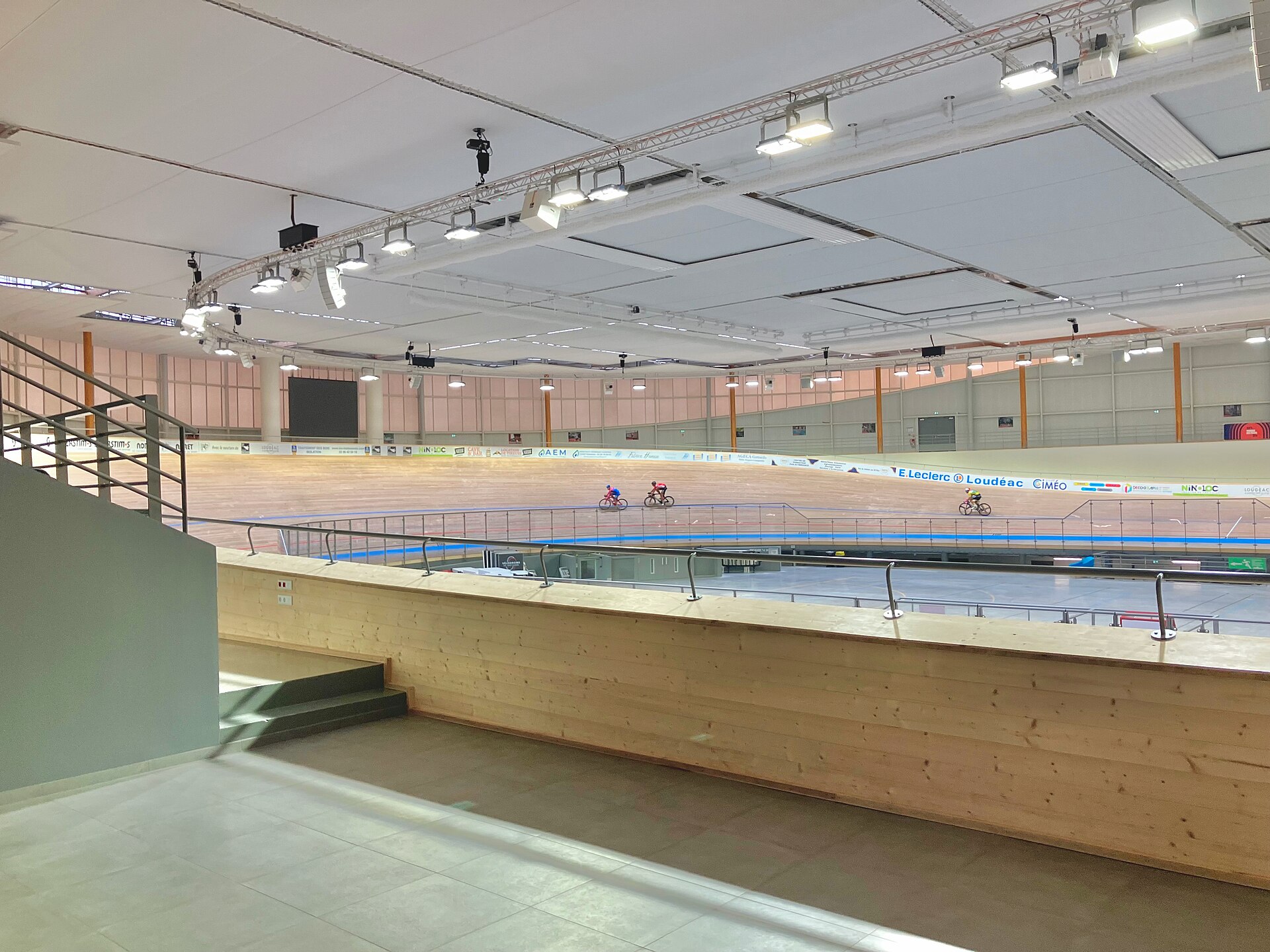 The interior of an indoor velodrome viewed from a walkway above the seating: a pale wooden banked track sweeping around a flat infield, a blue band along its inner edge and advertising boards above it.